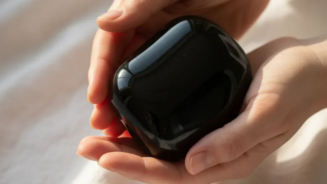 Manos femeninas sosteniendo una obsidiana negra brillante con luz natural lateral, piedra volcánica de protección y anclaje