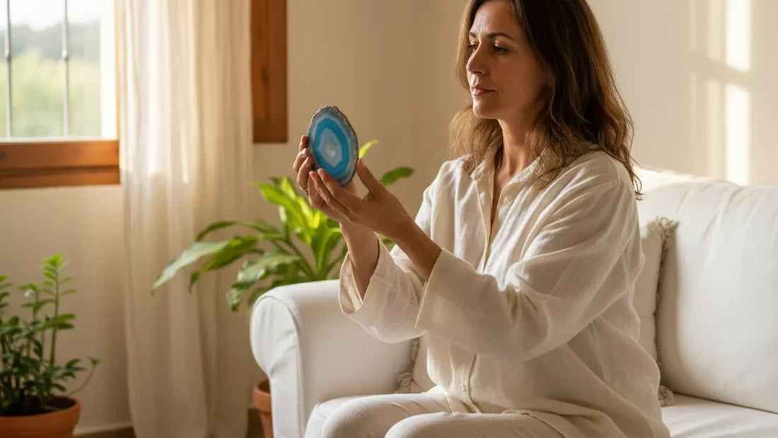 Mujer de 40 años sentada en sofá de lino blanco sosteniendo rodaja de ágata azul translúcida con ambas manos, luz natural de mañana, ambiente mediterráneo