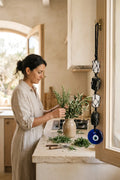 Amuleto de turmalina negra con piedras blancas de selenita y ojo turco azul colgado junto a una mujer preparando un ramo de hierbas en su cocina.