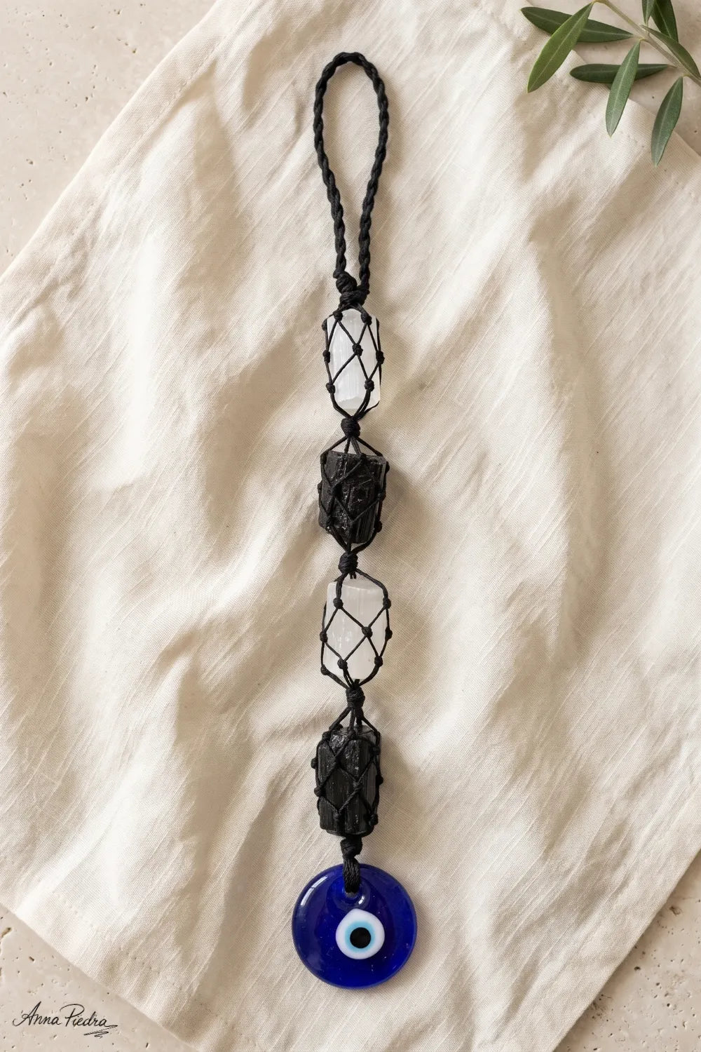 Amuleto de turmalina negra y selenita con ojo turco de cristal azul y cordón negro trenzado, presentado sobre tejido de lino beige.