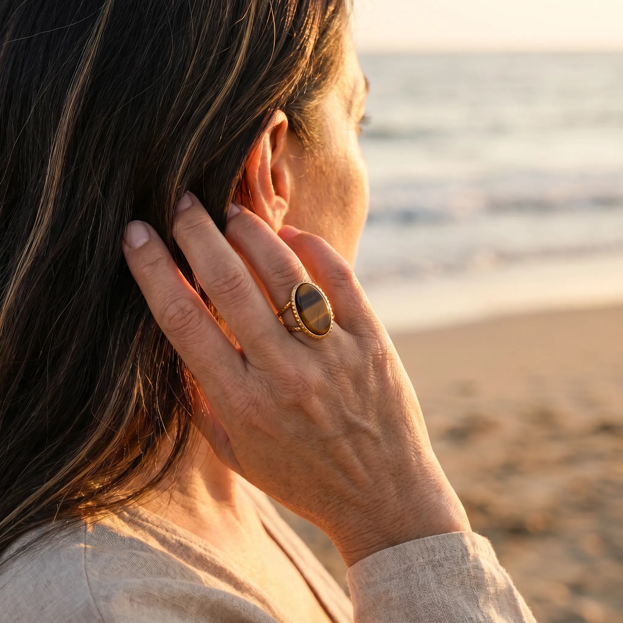 Mujer con anillo con ojo de tigre dorado llevando la mano al cabello frente al mar - Anna Piedra
