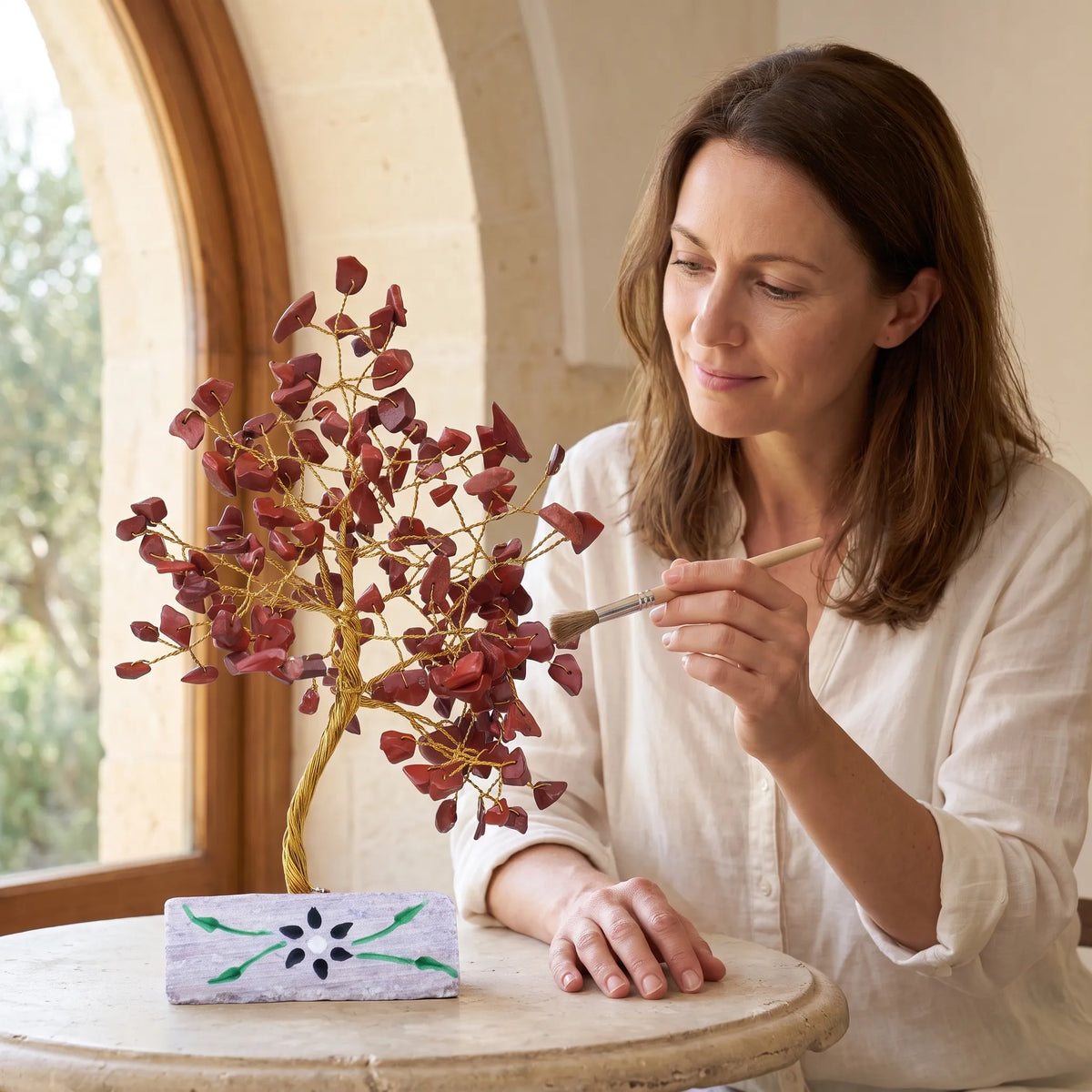 Mujer observando un Árbol de la Vida de jaspe rojo sobre una mesa redonda junto a una ventana con luz natural.