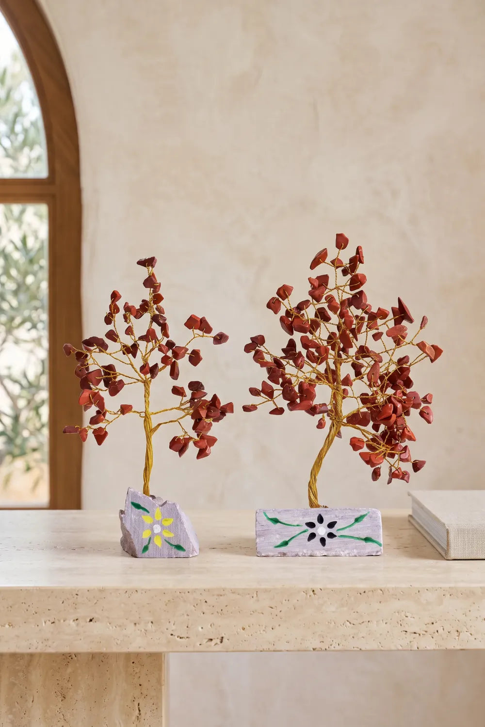 Dos Árboles de la Vida de jaspe rojo decorativos sobre una mesa de piedra en un interior luminoso.