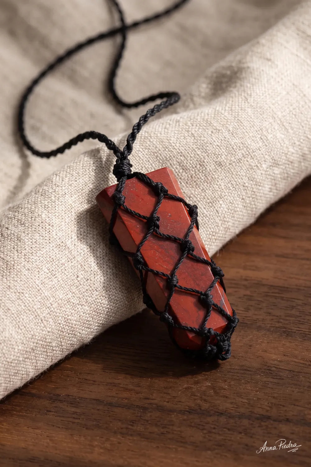 Detalle del colgante macramé de jaspe rojo sobre tela de lino beige y superficie de madera.