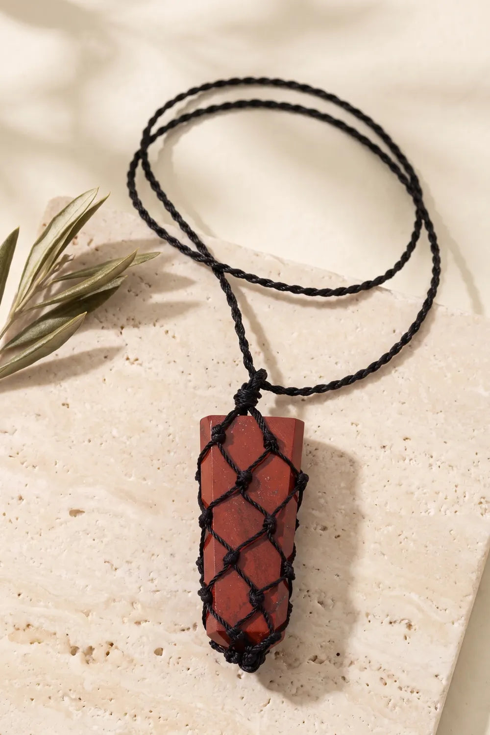 Detalle de un colgante macramé de jaspe rojo sostenido en la mano, con cordón negro ajustable.