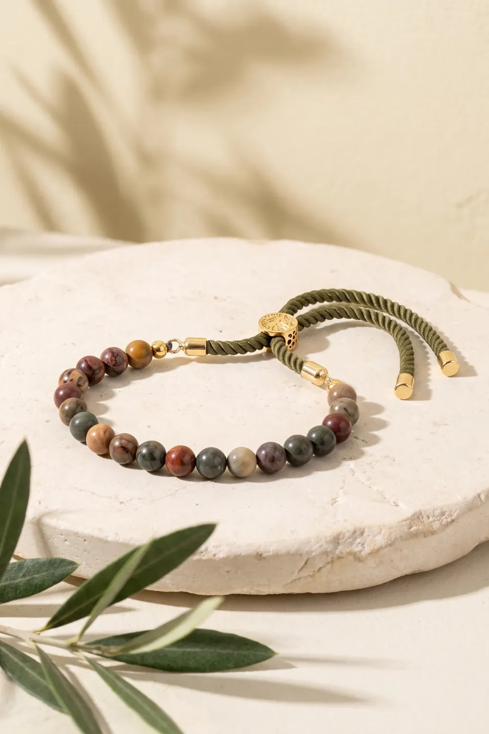 Pulsera de jaspe Picasso ajustable presentada sobre una base de piedra clara con ramas de olivo.