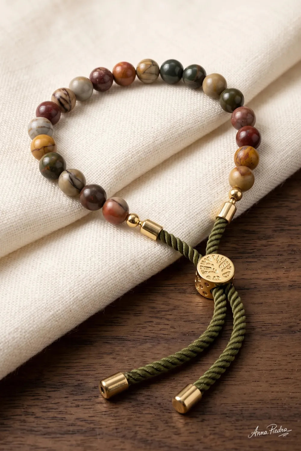 Detalle de una pulsera de jaspe Picasso ajustable con cordón verde y cierre dorado sobre tela de lino beige.
