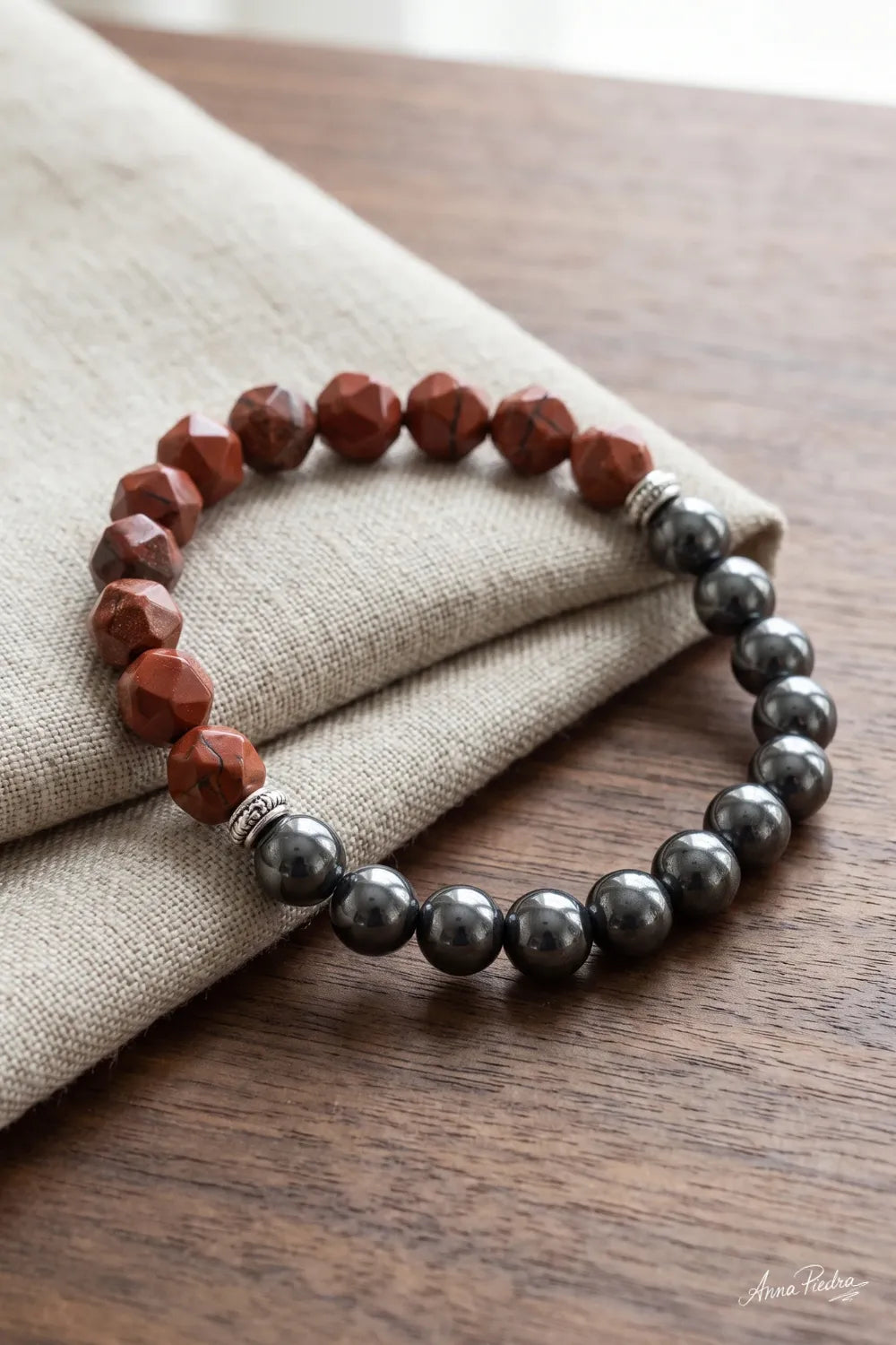 Detalle de una pulsera de jaspe rojo y hematita magnética sobre tela de lino beige y superficie de madera.