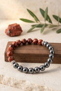Pulsera de jaspe rojo y hematita magnética presentada sobre un bloque de madera con una rama de olivo al fondo.