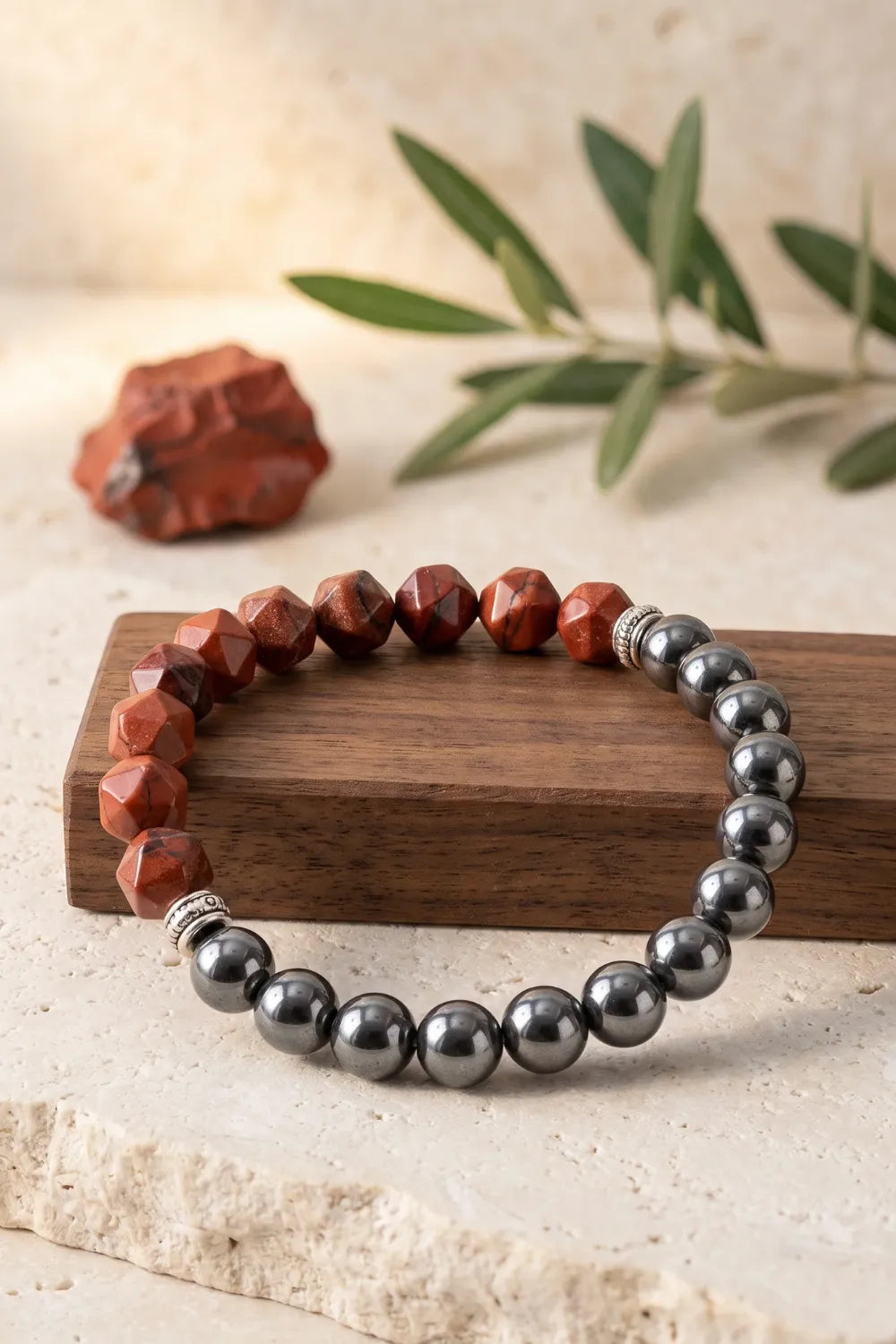 Pulsera de jaspe rojo y hematita magnética presentada sobre un bloque de madera con una rama de olivo al fondo.