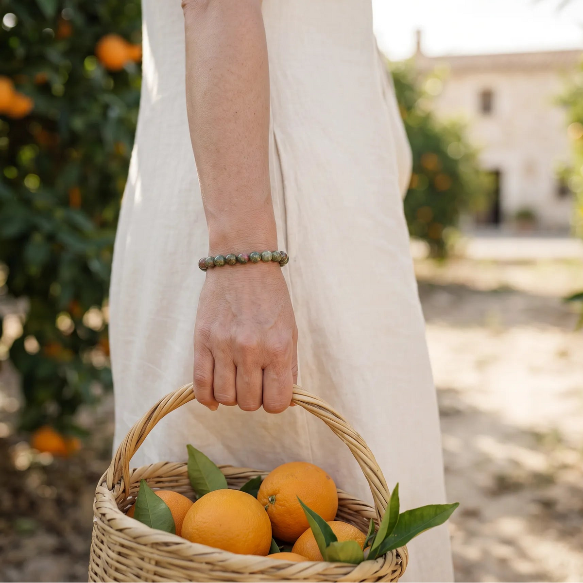 Mujer con pulsera de jaspe verde llevando una cesta con naranjas en un jardín mediterráneo.