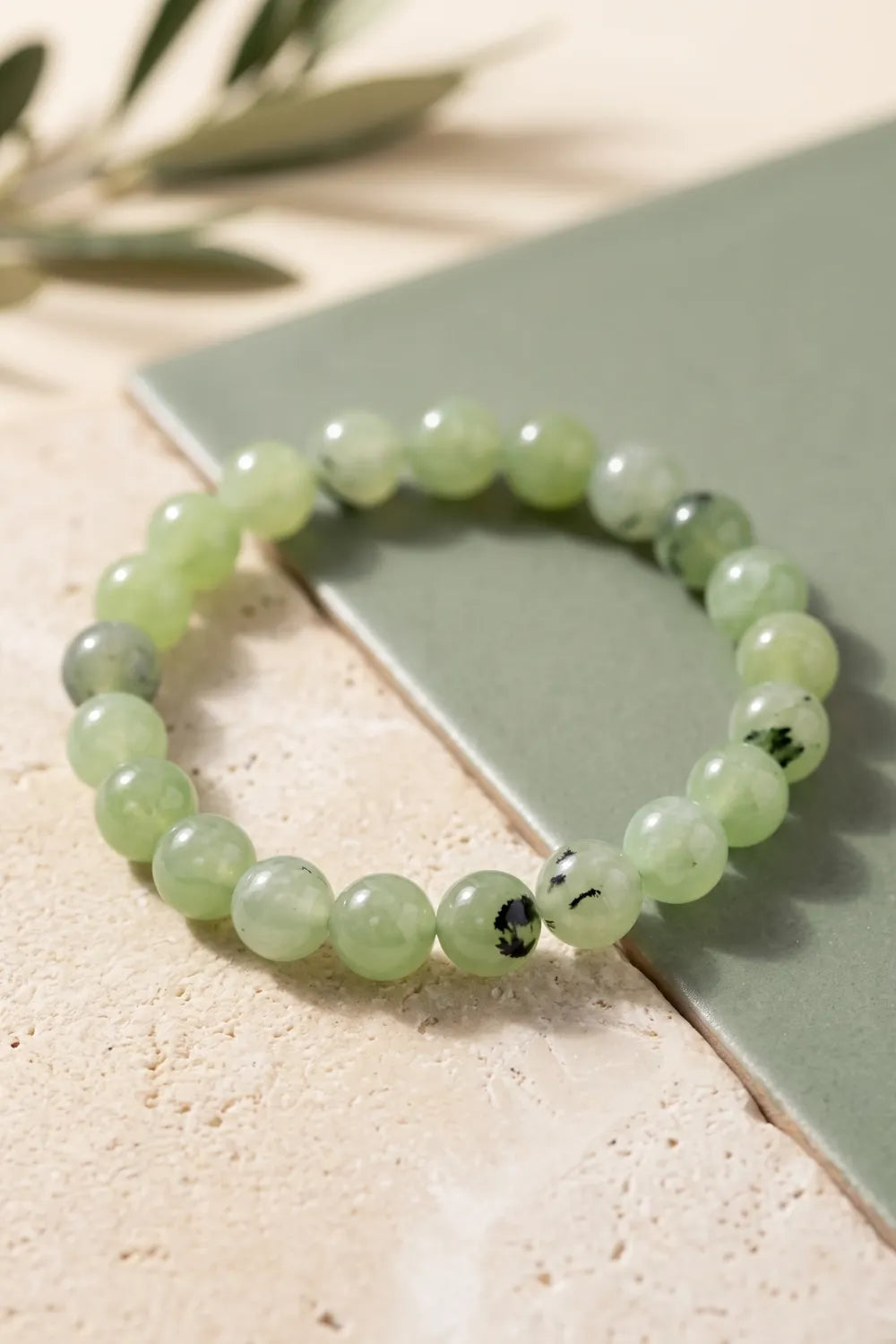 Primer plano de una pulsera de jade verde con cuentas redondas sobre superficie de piedra y cerámica verde.