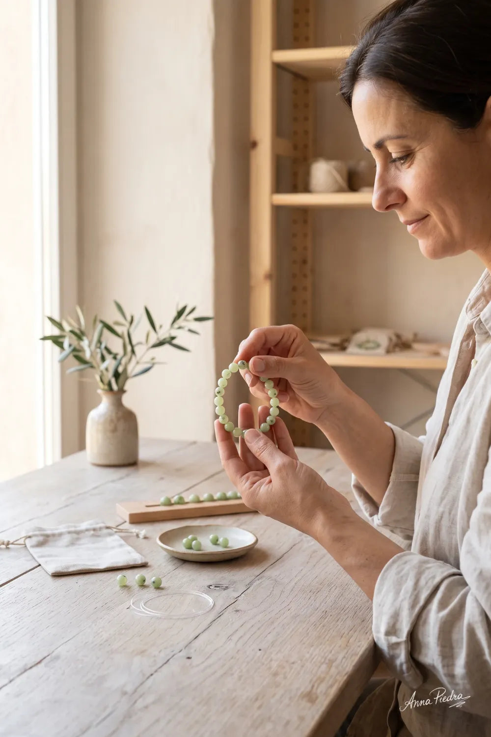 Mujer sosteniendo una pulsera de jade verde mientras trabaja en su creación sobre una mesa de madera.