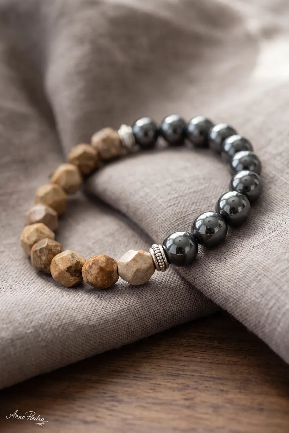 Primer plano de una pulsera de jaspe paisaje con cuentas beige facetadas, hematita magnética gris oscuro y detalles plateados sobre lino gris.