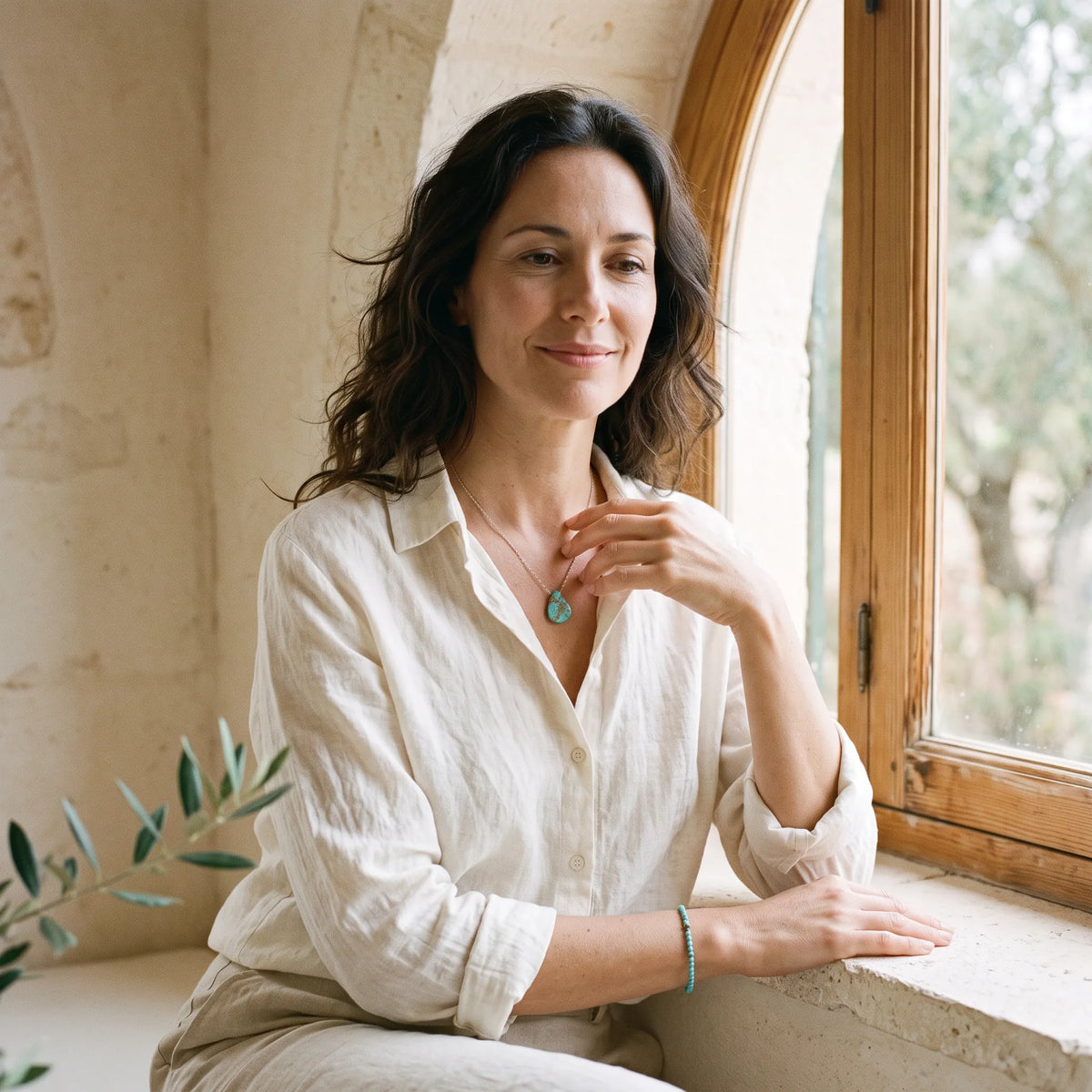 Mujer junto a una ventana con collar de turquesa natural y pulsera de cuentas azules en un ambiente mediterráneo luminoso.