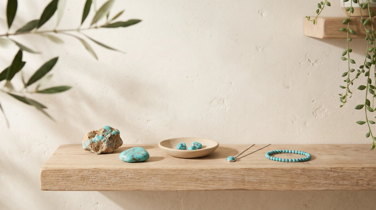 Colección de turquesa con piedra en bruto, cantos rodados, collar y pulsera de cuentas azules sobre una estantería de madera natural.