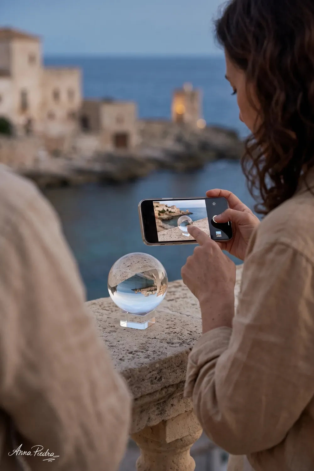 Mujer fotografiando con su móvil una bola de cristal transparente que refleja un paisaje costero mediterráneo.