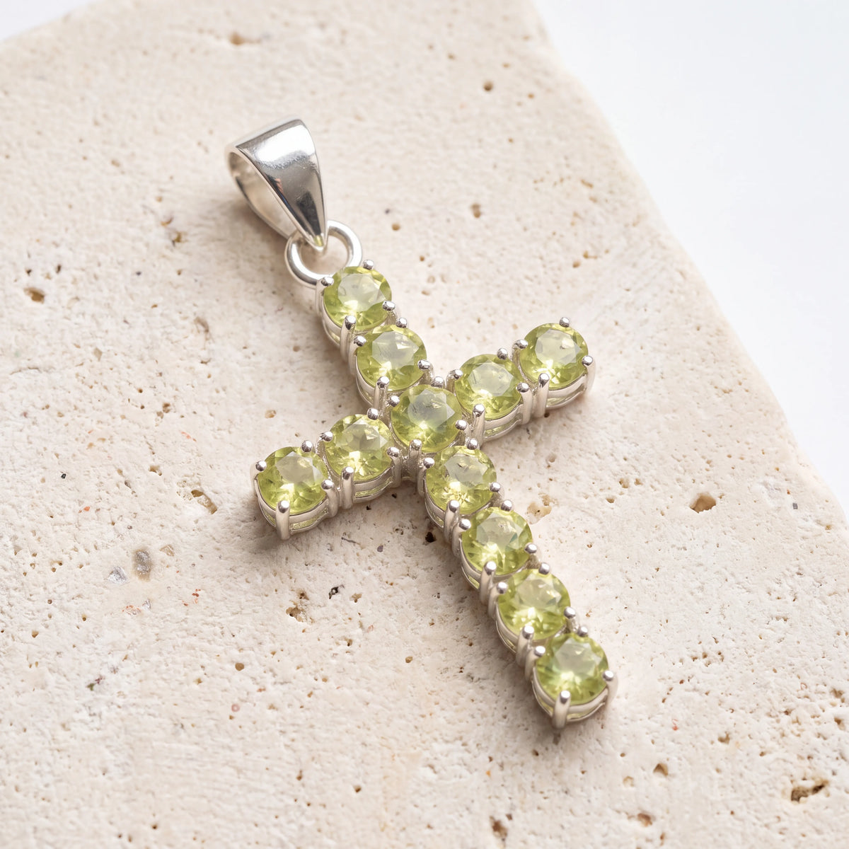 Colgante crucifijo de peridoto natural con montura de metal plateado sobre una superficie de piedra clara. anna piedra