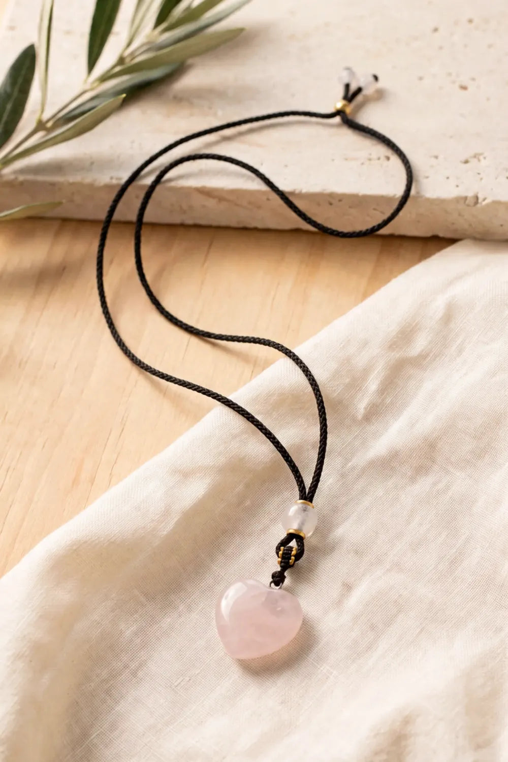 Colgante de cuarzo rosa en forma de corazón con cordón negro ajustable, sobre tela de lino beige y madera natural – Anna Piedra
 Copier