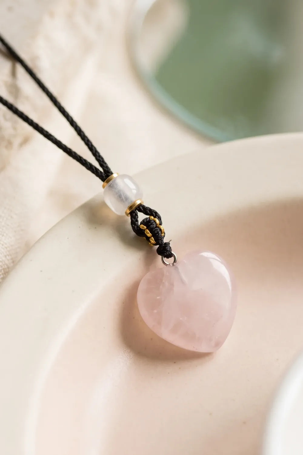 Primer plano de un colgante de cuarzo rosa natural en forma de corazón, con vetas rosadas, cordón negro trenzado y perla decorativa – Anna Piedra