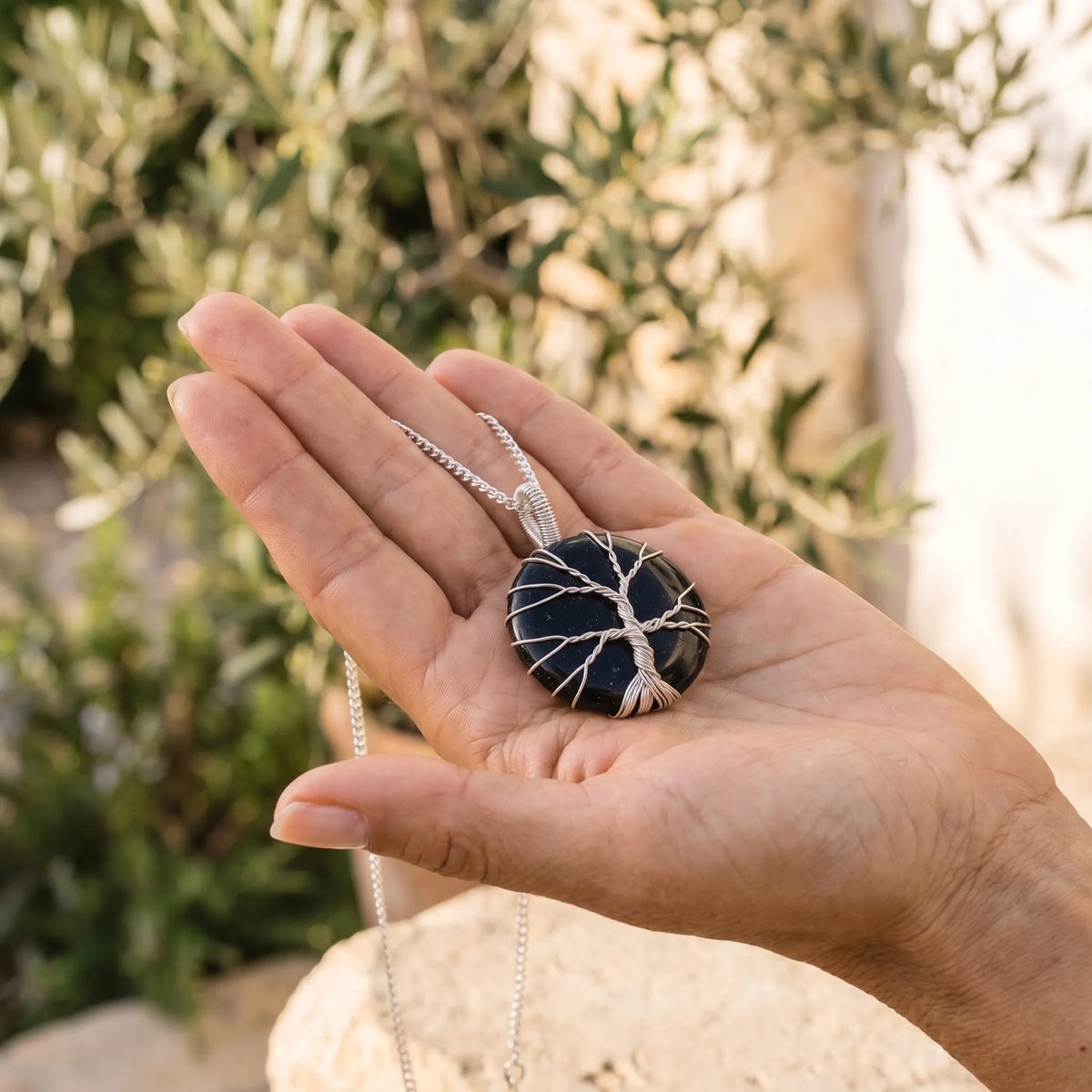 Mano sosteniendo un collar árbol de la vida de ónix negro con cadena plateada, fotografiado en un entorno exterior luminoso. anna piedra