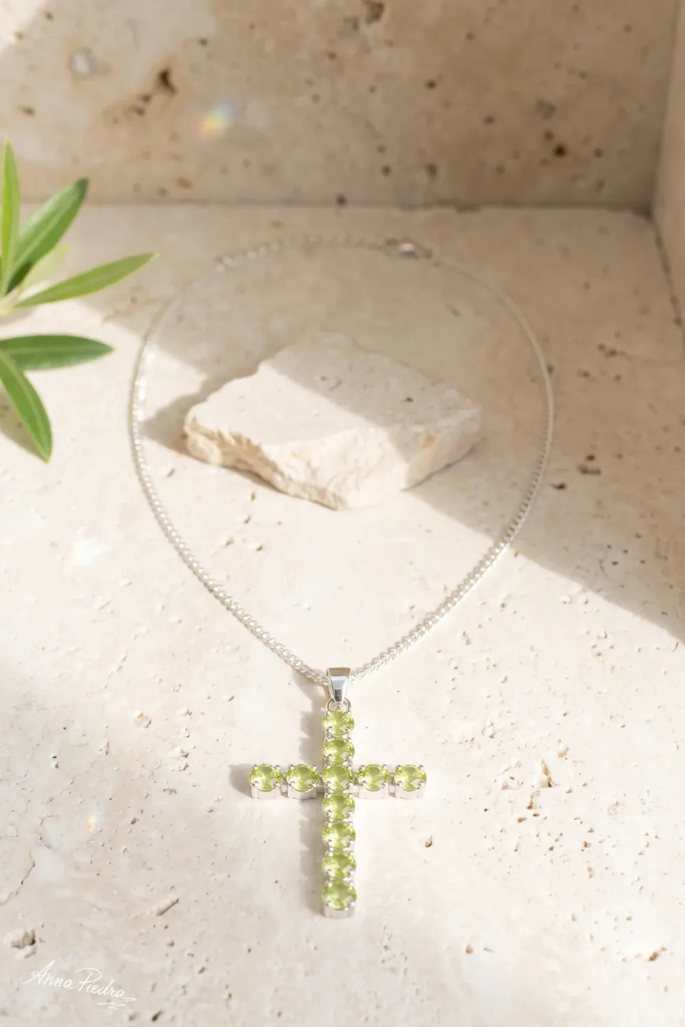 Collar crucifijo de peridoto natural con cadena plateada sobre una superficie de piedra clara.