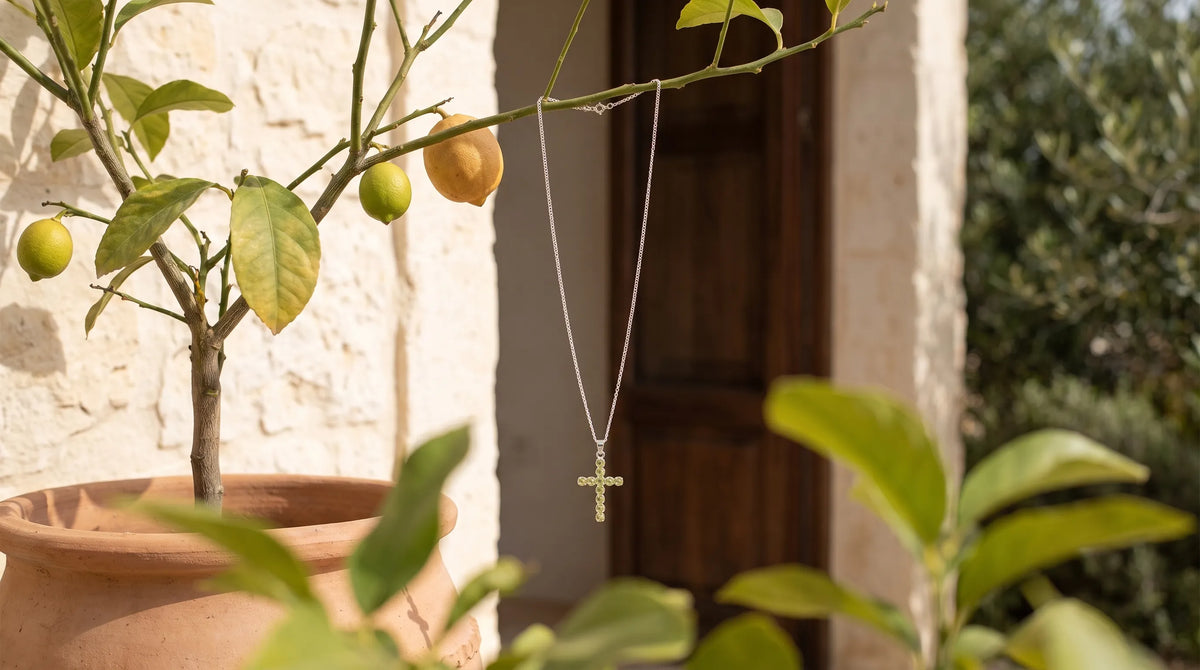 Collar crucifijo de peridoto con cadena plateada colgado de una rama de limonero en un entorno mediterráneo. anna piedra