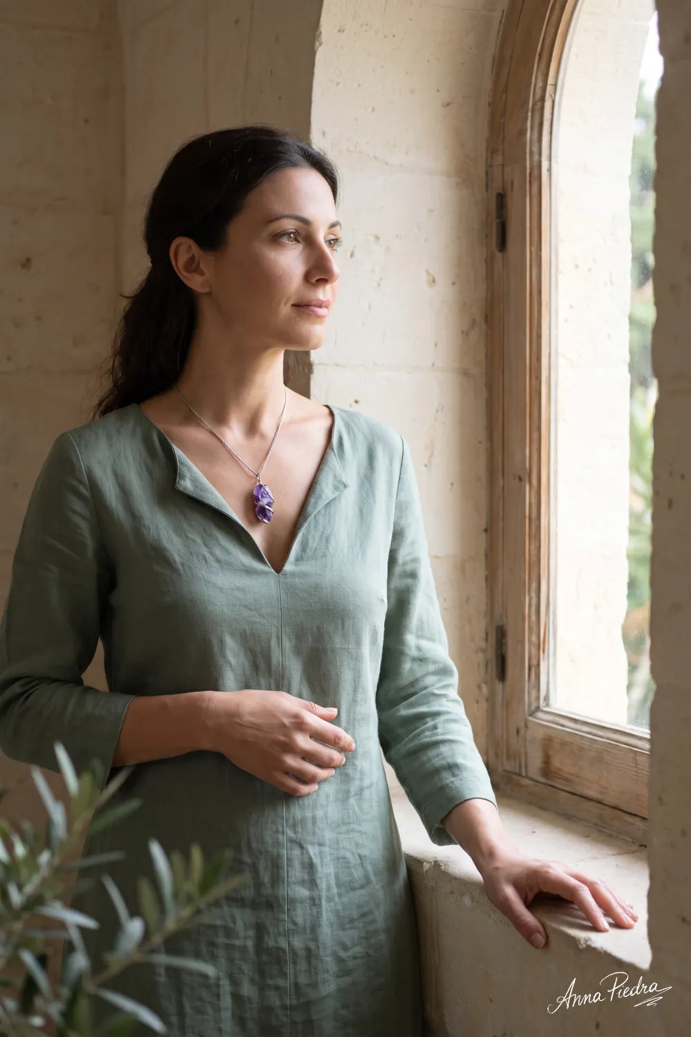 Mujer con collar de amatista en bruto y colgante violeta envuelto en alambre plateado, llevando una blusa verde salvia junto a una ventana – Anna Piedra