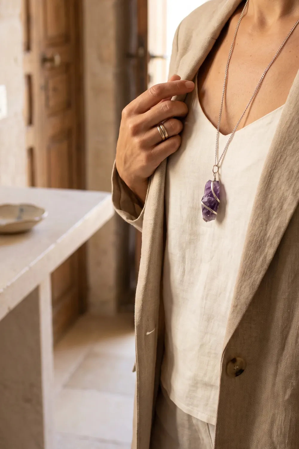 Detalle de collar de amatista en bruto con piedra violeta envuelta en metal plateado, llevado por una mujer con blusa blanca y chaqueta beige – Anna Piedra