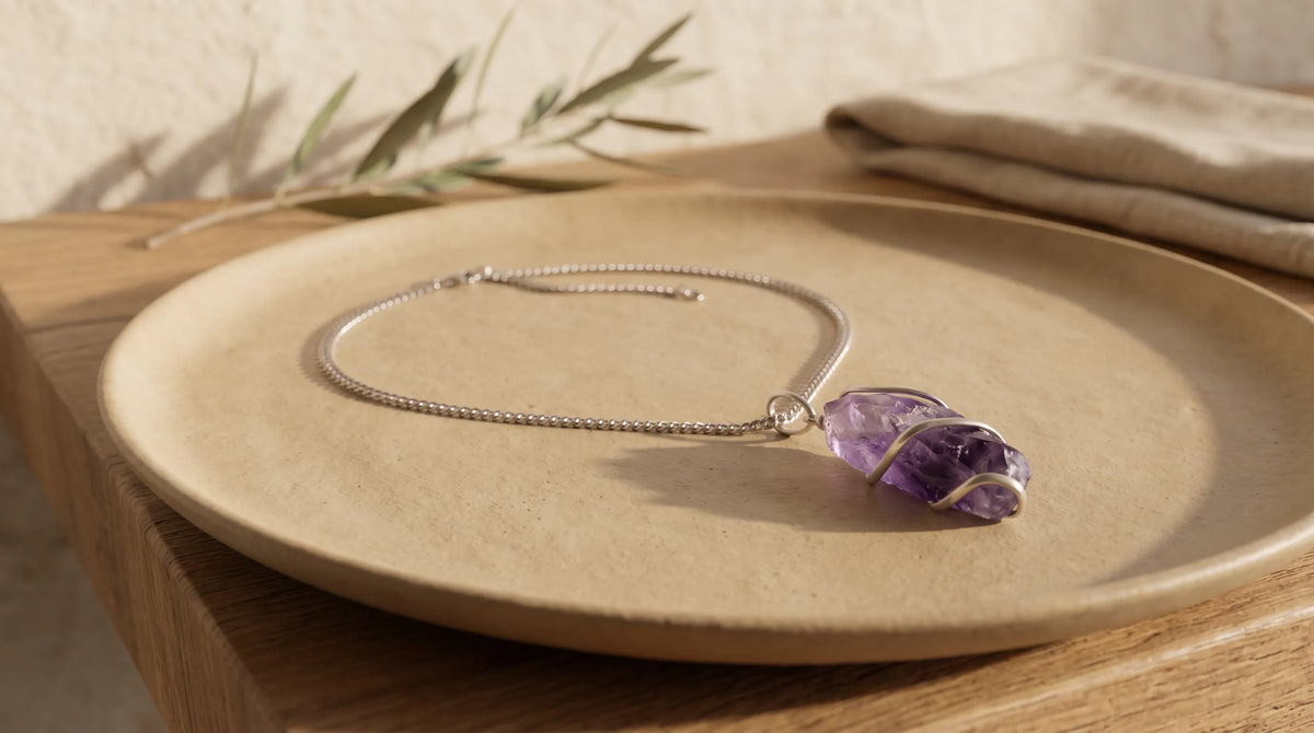 Collar de amatista en bruto con piedra violeta natural envuelta en alambre plateado y cadena metálica, sobre un plato de cerámica beige – Anna Piedra