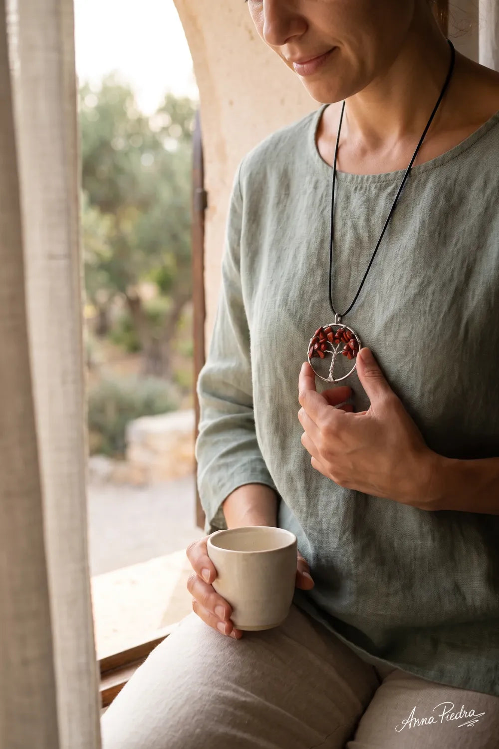 Mujer sujetando un collar de jaspe rojo con colgante Árbol de la Vida sobre una blusa verde salvia, junto a una ventana – Anna Piedra