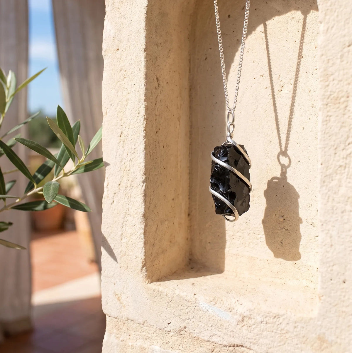 Collar de ónix negro en bruto con cadena plateada colgado en una pared de piedra clara con luz natural.