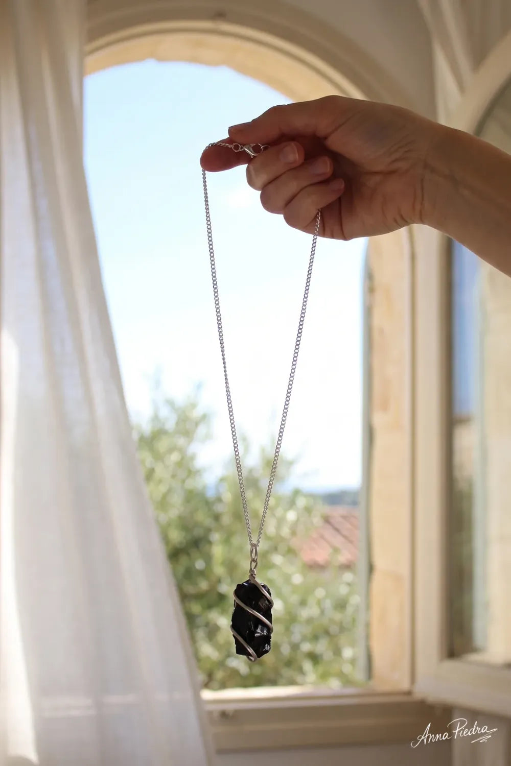 Mano sosteniendo un collar de ónix negro en bruto con cadena plateada frente a una ventana luminosa. anna piedra