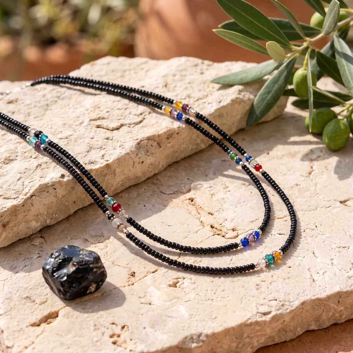 Collar nepalí de ónix negro de doble hilera con pequeñas cuentas de colores colocado sobre piedra clara junto a una piedra de ónix. anna piedra