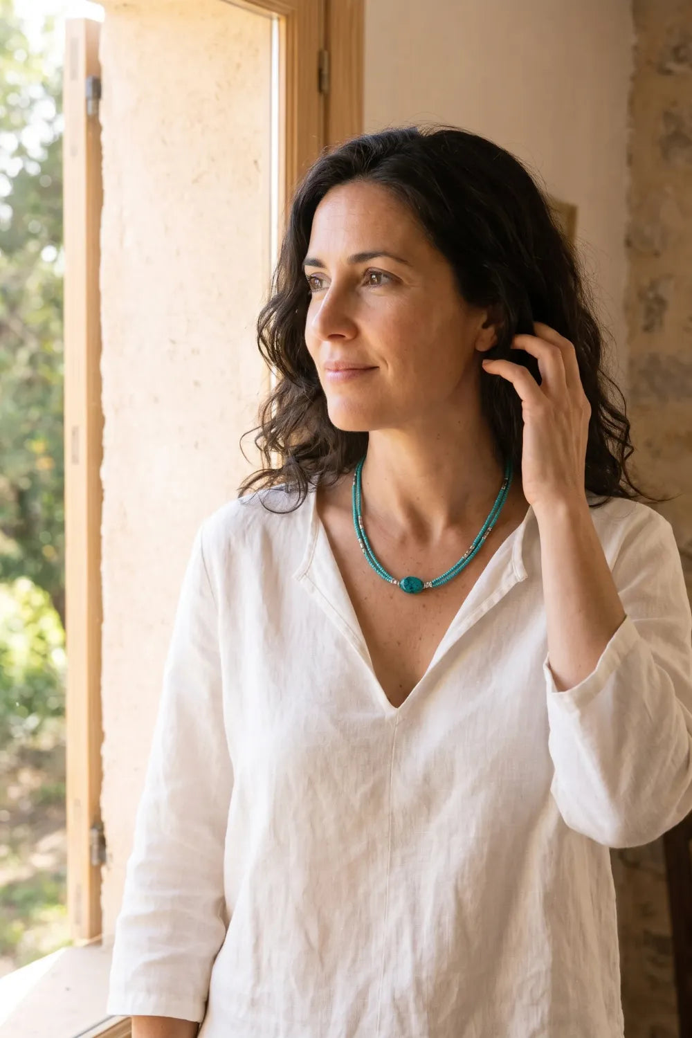 Mujer con camisa blanca llevando un collar de turquesa de dos vueltas junto a una ventana de madera con luz natural.