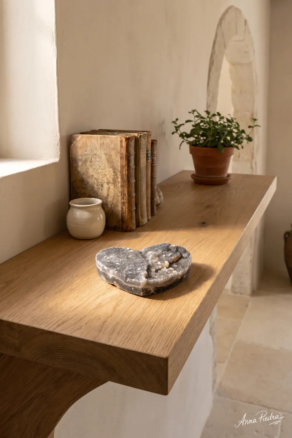 Corazón de calcita negra colocado sobre una consola de madera junto a libros y una planta en un interior luminoso. anna piedra