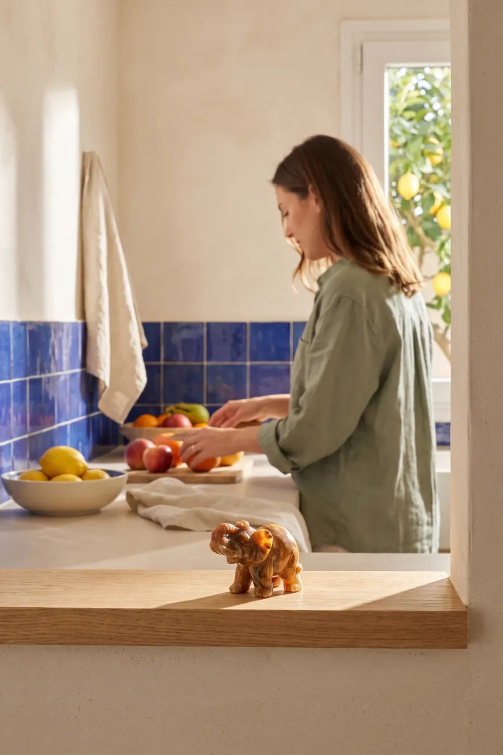 Elefante de la suerte de ágata encaje loco en el alféizar de una cocina, con mujer preparando fruta al fondo - Anna Piedra