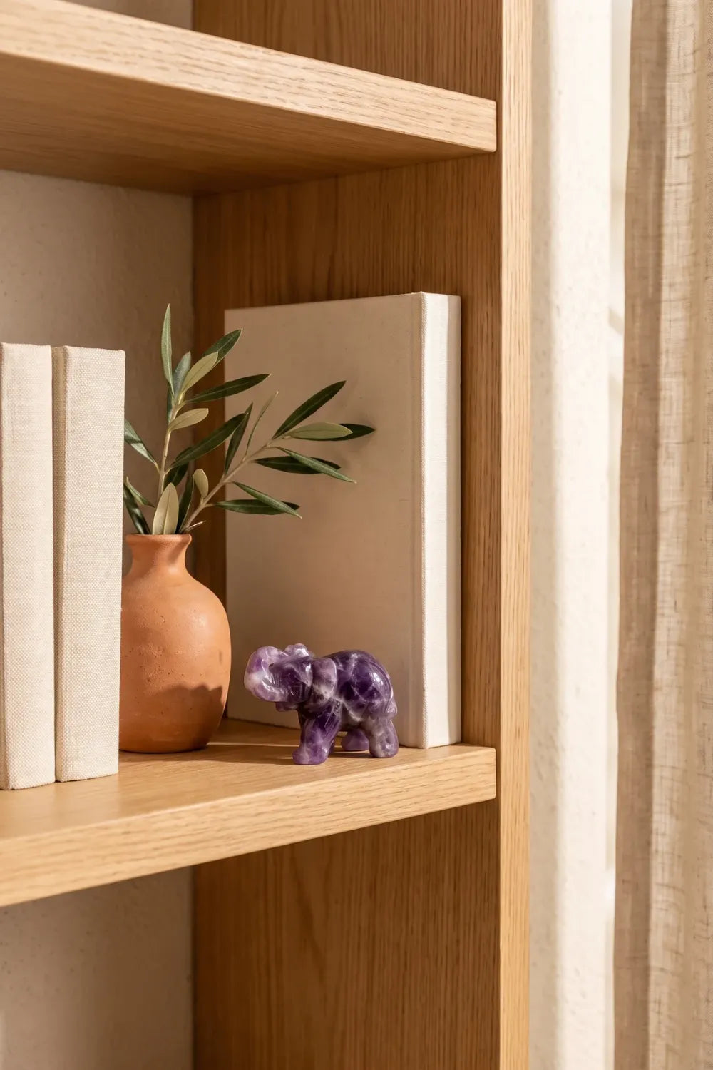 Elefante de la suerte de amatista sobre una estantería de madera junto a libros y un jarrón con olivo - Anna Piedra