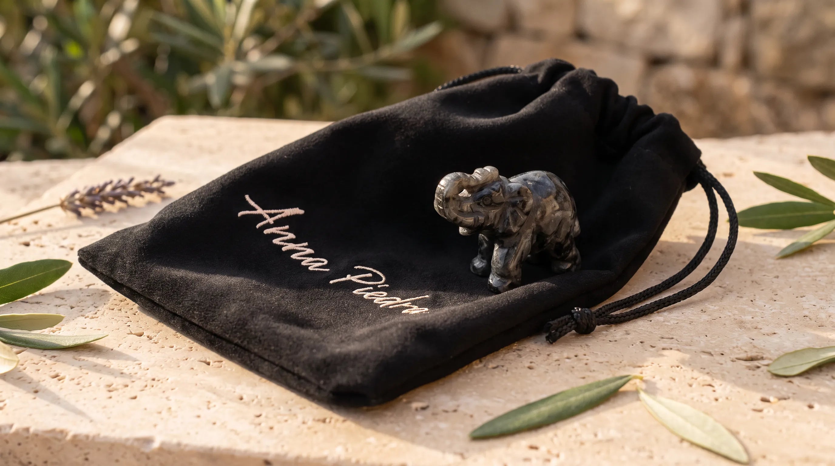 Elefante de la suerte tallado en labradorita sobre una bolsa de terciopelo Anna Piedra, en una terraza con olivo - Anna Piedra