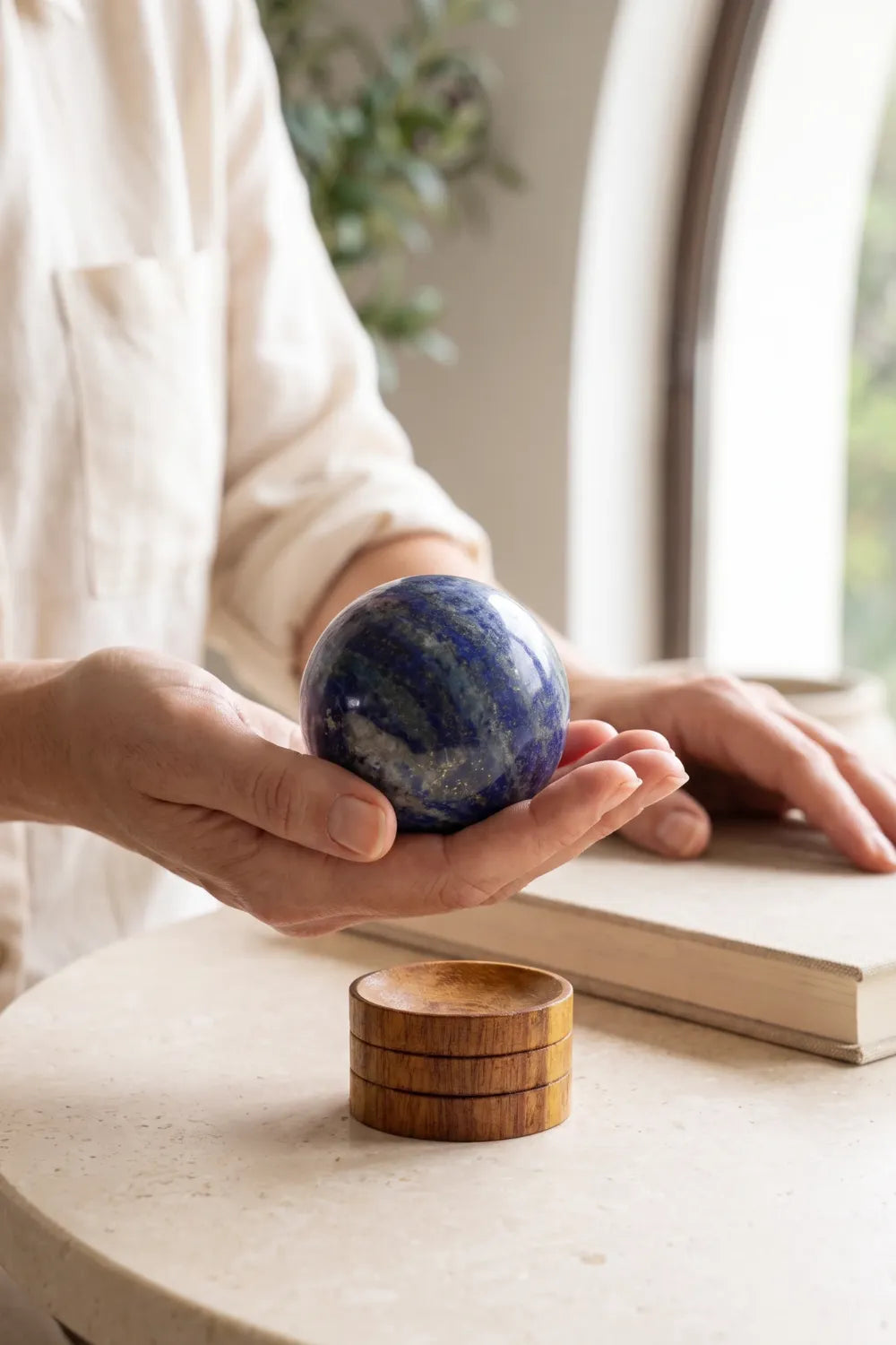 Mano sosteniendo una esfera de lapislázuli azul pulida junto a un libro sobre una mesa clara