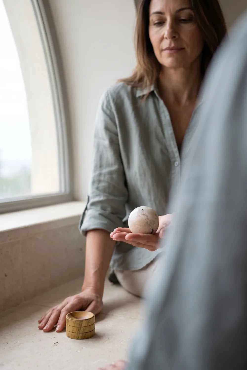 Mujer sosteniendo una esfera de piedra luna de color blanco con vetas grises, junto a una ventana y un soporte de madera – Anna Piedra