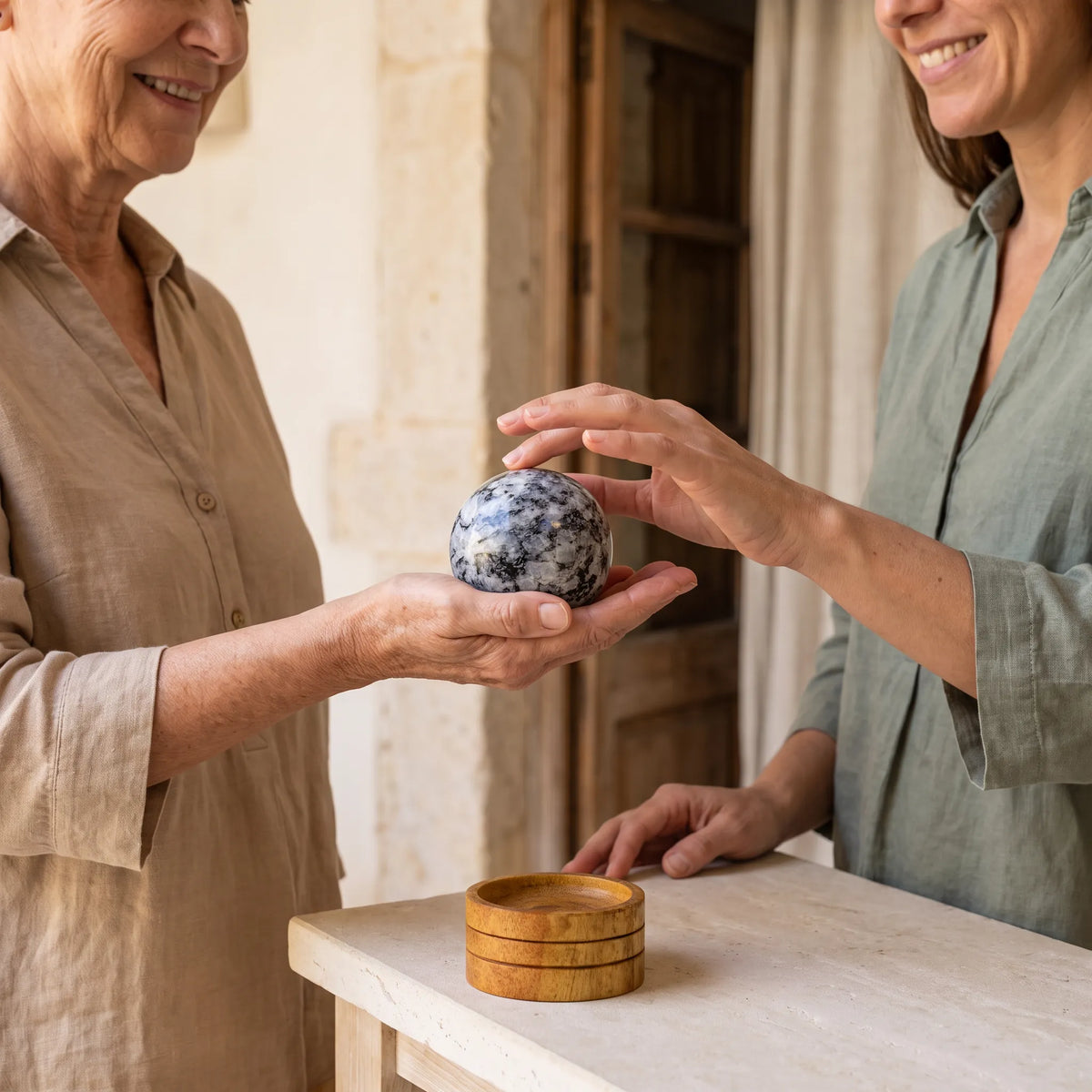 Esfera de piedra luna pulida con vetas blancas y negras, sostenida entre las manos de dos mujeres junto a una base de madera – Anna Piedra