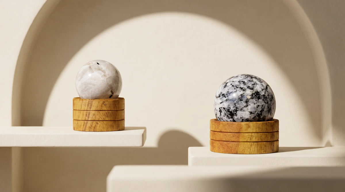 Dos esferas de piedra luna natural de tonos blancos, grises y negros, sobre bases de madera en estantes de decoración beige – Anna Piedra