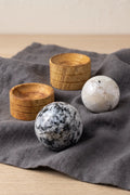 Dos esferas de piedra luna pulida con vetas naturales blancas y negras, junto a sus soportes de madera sobre una tela gris – Anna Piedra