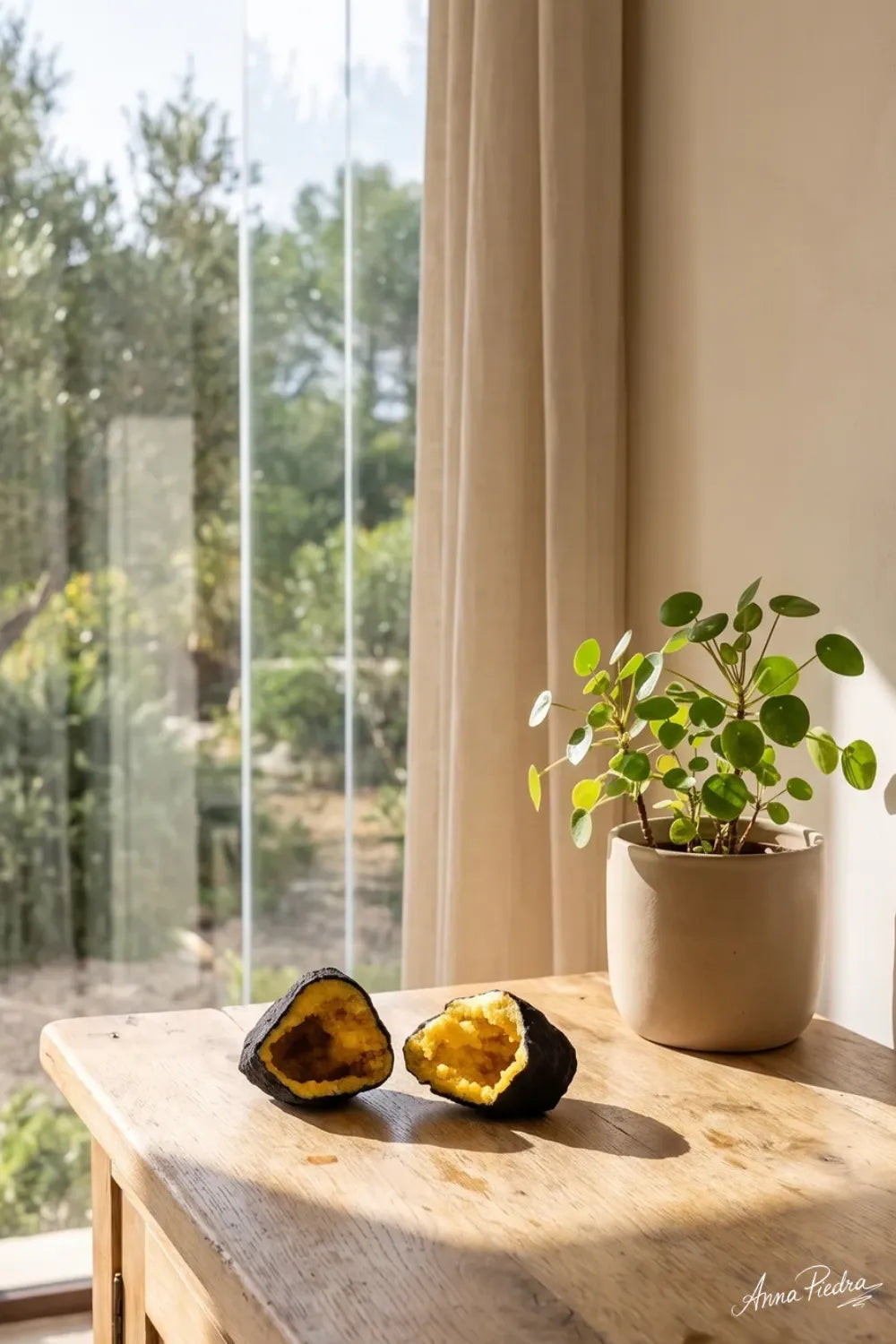 Dos mitades de una geoda de calcita amarilla sobre un mueble de madera junto a una ventana y una planta verde. anna piedra