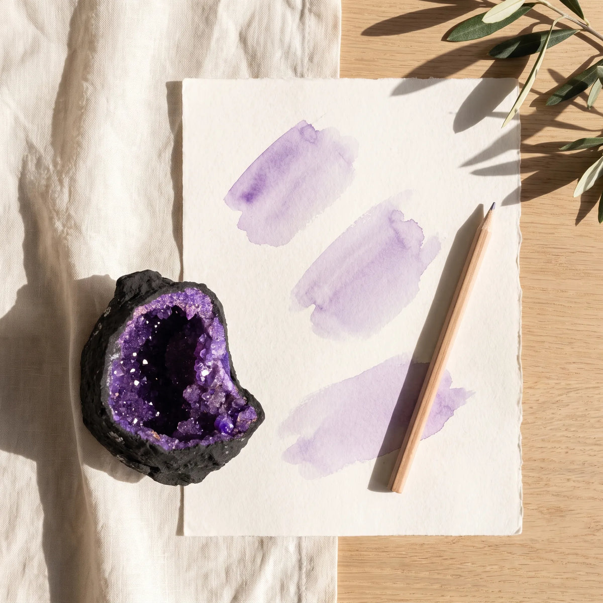 Geoda de calcita morada junto a una hoja con pinceladas de acuarela lila y un lápiz sobre una mesa de madera clara. anna piedra