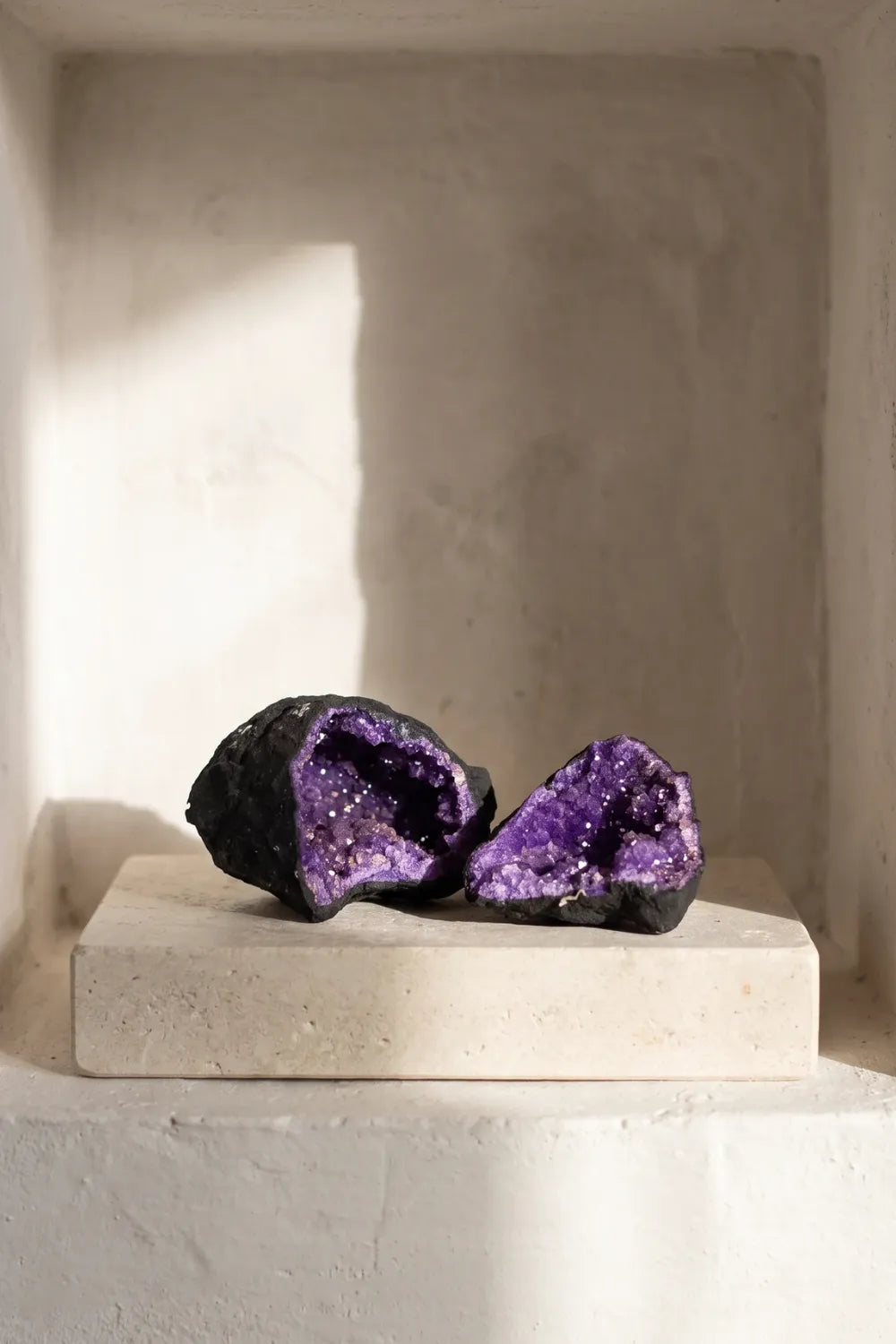 Dos mitades de una geoda de calcita morada expuestas sobre una base clara en un interior luminoso. anna piedra