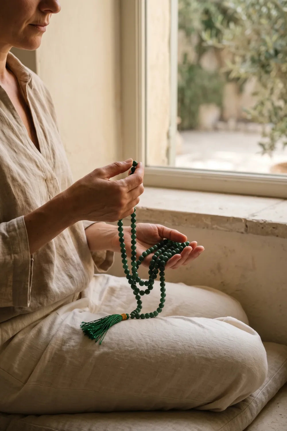 Mujer sentada junto a una ventana pasando las cuentas de un japamala de jade verde durante un momento de meditación.