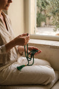 Mujer sentada junto a una ventana pasando las cuentas de un japamala de jade verde durante un momento de meditación.