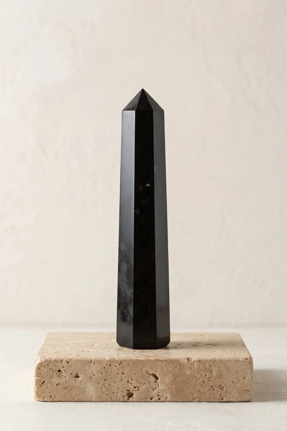 Obelisco de turmalina negra pulida presentado en vertical sobre una base de piedra beige con fondo neutro.